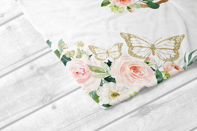 K&ouml;nnte beinhalten: Ein wei&szlig;er Stoff mit einem floralen Muster aus rosa Rosen, gr&uuml;nen Bl&auml;ttern und goldenen Glitzer-Schmetterlingen. Der Stoff ist &uuml;ber eine wei&szlig;e Holzoberfl&auml;che gefaltet.
