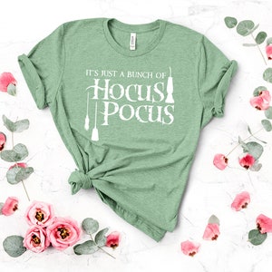 May include: A green t-shirt with the text "It's just a bunch of Hocus Pocus" in white lettering. The shirt is folded and displayed on a white background with pink flowers and green leaves.