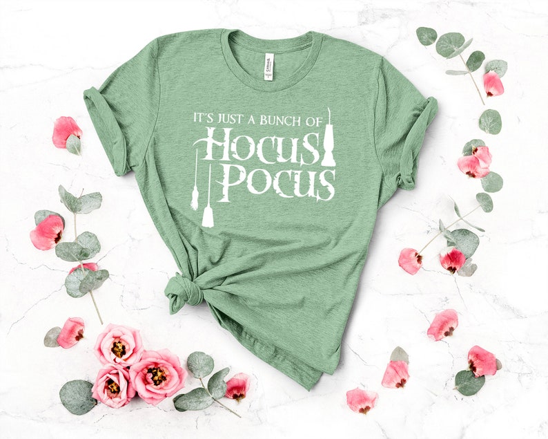 May include: A green t-shirt with the text "It's just a bunch of Hocus Pocus" in white lettering. The shirt is folded and displayed on a white background with pink flowers and green leaves.