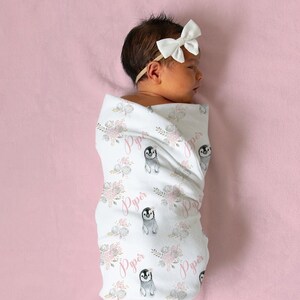 May include: A white swaddle blanket with a pink floral and penguin pattern. The blanket has the name "Piper" printed on it multiple times. A baby is wrapped in the blanket.
