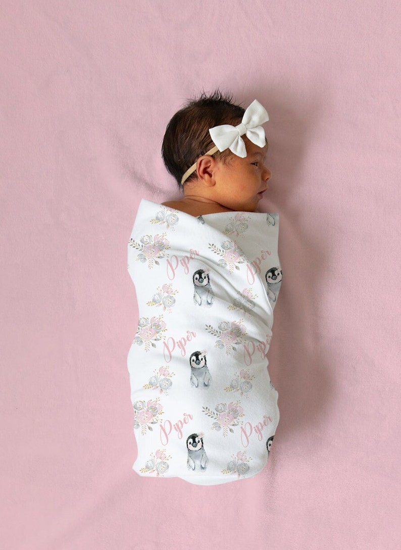 May include: A white swaddle blanket with a pink floral and penguin pattern. The blanket has the name "Piper" printed on it multiple times. A baby is wrapped in the blanket.