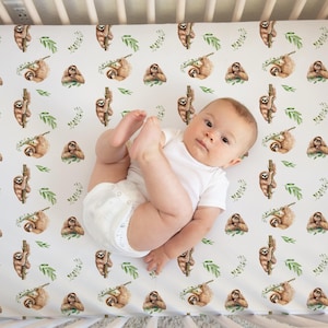 May include: A baby in a white onesie lies on a crib sheet with a repeating pattern of watercolor sloths and green leaves on a white background. The crib sheet is in a crib with white wooden slats.
