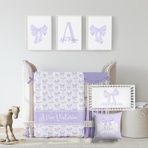 May include: A lavender and white nursery set, including a cot, bedding, and wall art. The set features a cot bumper, blanket, and pillow, all decorated with bow patterns. The wall art includes bow and initial prints, with the name "Alice Victoria".
