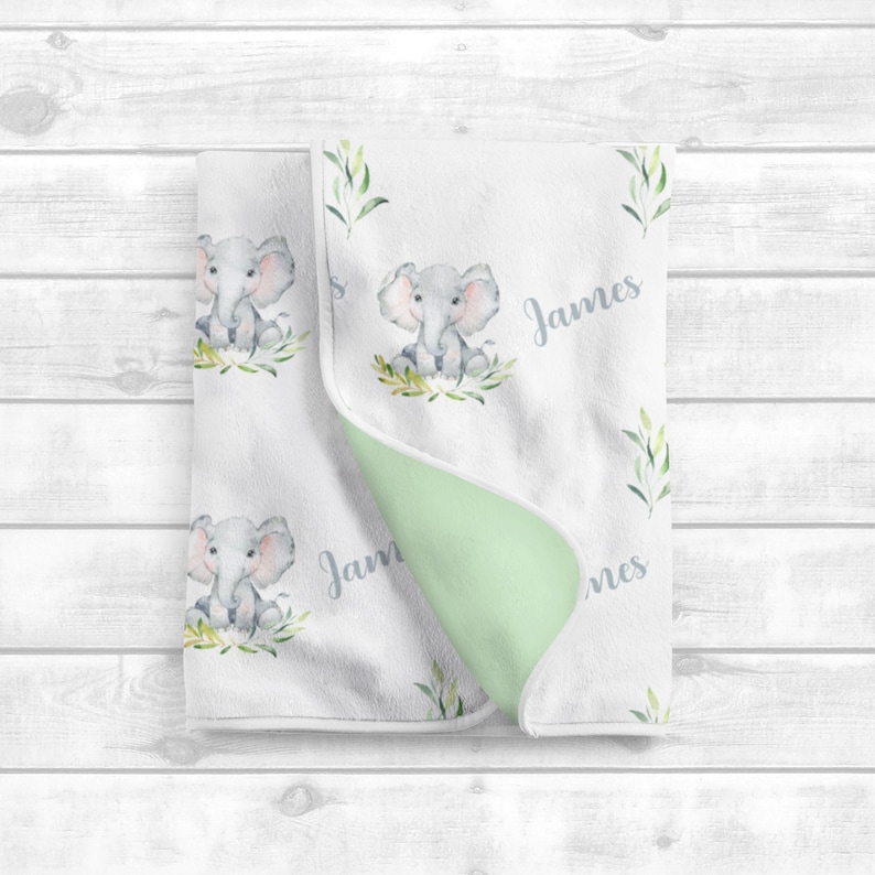 May include: A white baby blanket with a green and white elephant print. The blanket is personalized with the name "James".