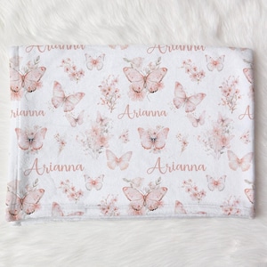 Puede incluir: Manta de bebé blanca con un estampado repetido de mariposas rosas, flores y el nombre "Arianna" en escritura cursiva. La manta es suave y acogedora, perfecta para una habitación infantil.