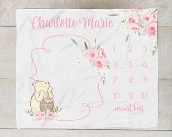 May include: White baby milestone blanket with pink floral accents and the name "Charlotte Marie". Features a Winnie the Pooh illustration and a monthly tracker from 1 to 12 months. A perfect baby shower gift.
