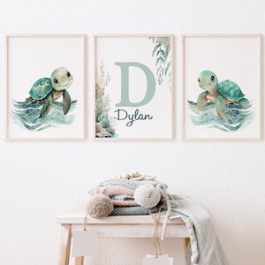 May include: Three framed watercolour prints. Two feature a teal and green sea turtle in blue water. The central print has a large teal letter "D" and the name "Dylan". The prints are in light wood frames, ideal for a nursery or child's room.