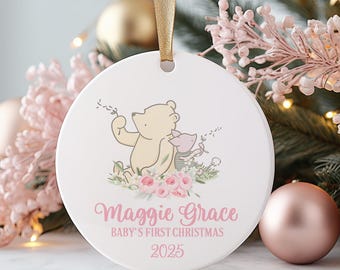 Op de afbeelding: Een witte keramische kerstbal met een gouden lint. De ornamenten hebben een Winnie de Poeh en Knorretje illustratie met roze bloemaccenten. Tekst luidt "Maggie Grace, Baby's First Christmas, 2025."