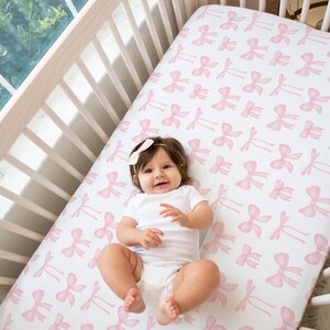 May include: A baby in a white onesie and a white bow headband lies on a crib sheet with a pink bow pattern. The crib is made of light-colored wood.