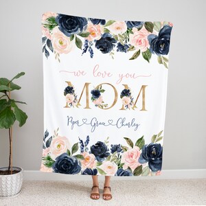 May include: A white blanket with a floral design in shades of navy blue and pink. The blanket features the text "we love you MOM" in gold lettering with a pink heart and the names "Piper, Grace, Charley" in cursive writing below.