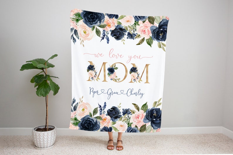 May include: A white blanket with a floral design in shades of navy blue and pink. The blanket features the text "we love you MOM" in gold lettering with a pink heart and the names "Piper, Grace, Charley" in cursive writing below.