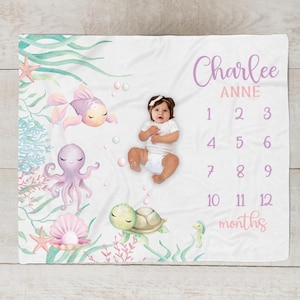 May include: A white baby milestone blanket with an underwater theme. The blanket features illustrations of a fish, octopus, turtle, and seahorse. The baby's name is written in cursive, with numbers 1-12 to track the baby's age in months.