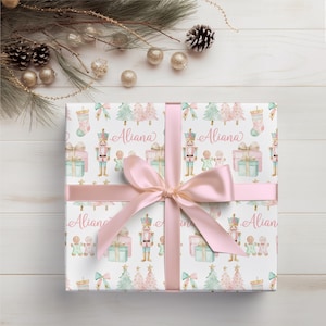 Op de afbeelding: Een cadeau verpakt met een wit papier met een herhaald patroon van kerstbomen, notenkrakers, peperkoekenmannetjes en cadeau dozen in pastelkleuren. Het cadeau is vastgebonden met een roze lint en strik. De naam "Aliana" staat op het cadeaupapier.