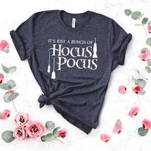 May include: A navy blue t-shirt with the text "It's just a bunch of Hocus Pocus" in white. The text is accompanied by a graphic of three broomsticks and a cauldron.