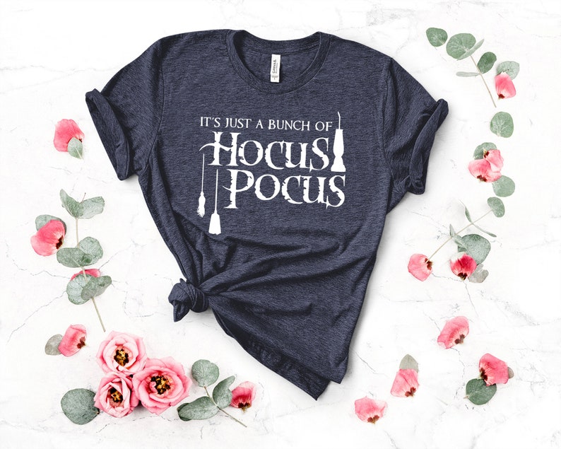 May include: A navy blue t-shirt with the text "It's just a bunch of Hocus Pocus" in white. The text is accompanied by a graphic of three broomsticks and a cauldron.