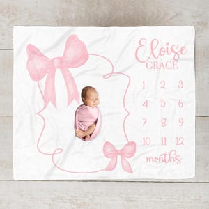 May include: White baby milestone blanket with pink bow illustrations and the name "Eloise Grace". The blanket features a space for a baby photo and numbers 1-12 for monthly milestones.
