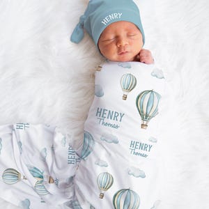 May include: A newborn baby wrapped in a white swaddle blanket and matching cap. The blanket and cap are light blue and feature a hot air balloon design with the name "Henry" printed on them. The baby is lying on a white, fluffy surface.