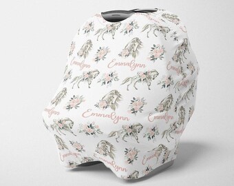 May include: White baby car seat cover with a repeating pattern of gray horses, pink flowers, and the name "Emmalynn" in a cursive font. The cover is designed to fit over a car seat.