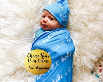 May include: A baby wrapped in a blue swaddle blanket and matching hat, both printed with the name "Luke" in white script. The baby is lying on a fluffy white surface. The image includes the text "Choose Your Own Colors" and "Free Shipping".