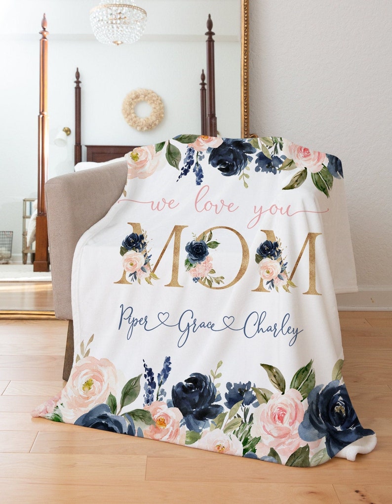 May include: A white fleece blanket with a floral design in shades of pink and navy blue. The blanket features the words "we love you MOM" in gold lettering and the names "Piper, Grace, Charley" in cursive script with hearts.