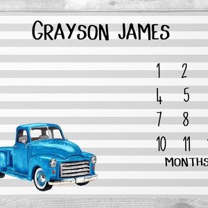 May include: A baby milestone blanket with a blue vintage truck illustration. The blanket features the name "Grayson James" and numbers 1-12 with the word "Months". The background has gray and white stripes.