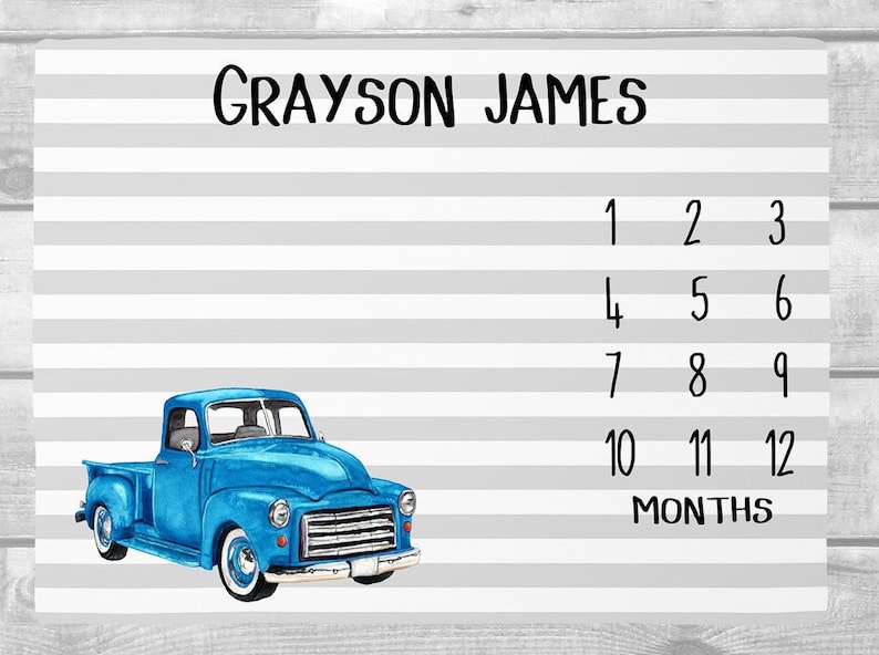 May include: A baby milestone blanket with a blue vintage truck illustration. The blanket features the name "Grayson James" and numbers 1-12 with the word "Months". The background has gray and white stripes.