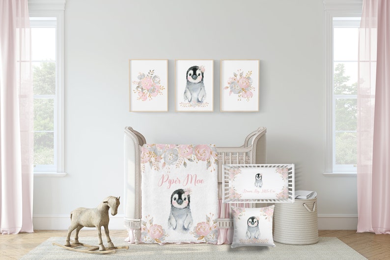 May include: A nursery bedding set with a pink and gray color scheme featuring a penguin with a floral crown. The set includes a blanket, a pillow, and a crib sheet. The blanket and pillow feature a floral design with gold accents. The crib sheet has a floral border and the text "Dream Big Little One".
