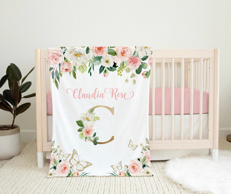 K&ouml;nnte beinhalten: Eine wei&szlig;e Babydecke mit einem Blumendesign in Rosa- und Gr&uuml;nt&ouml;nen. Die Decke zeigt einen gro&szlig;en goldenen Buchstaben "C", der von Blumen und Schmetterlingen umgeben ist. Der Text "Claudia Rose" ist in rosa Kursivschrift geschrieben.