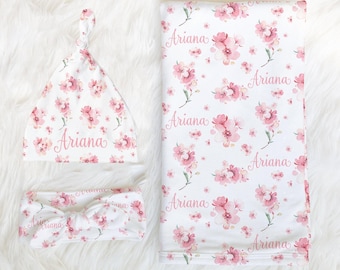 May include: A white baby hat with a top knot, a matching knotted headband, and a rectangular blanket, all featuring a repeating pink floral pattern and the name Ariana printed in a cursive script across the fabric surfaces.