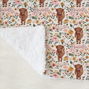 May include: A soft blanket with a floral pattern featuring brown Highland cows and the name "Danielle" in pink script. The blanket has a white sherpa backing. The blanket is made of soft material.