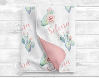 May include: A white and pink baby blanket with a cactus pattern and the name "Selena" written in pink. The blanket is folded in half, showing the pink side.