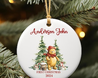 Op de afbeelding: Witte kerstbal van keramiek met een cartoonbeer die een rode muts en sjaal draagt. De kerstbal heeft de tekst "Anderson John" en "First Christmas 2024" in rood.