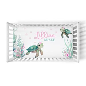 May include: A white crib sheet featuring watercolor illustrations of sea turtles and aquatic plants. The sheet includes the name "Lillian Grace" in pink and teal lettering, with pink polka dots scattered across the design.