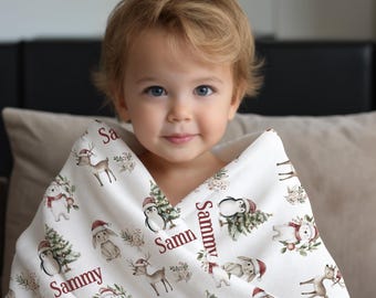 May include: A white baby blanket with a festive Christmas design, including reindeer, penguins, snowmen, and bunnies in Santa hats. The name "Sammy" is printed in burgundy. The blanket is wrapped around a young child.