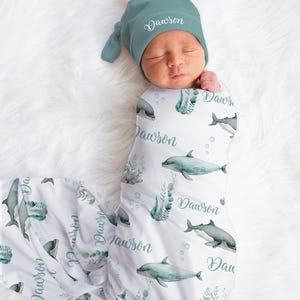 Puede incluir: Un bebé recién nacido envuelto en una manta blanca y un gorro a juego. La manta presenta un estampado de delfines y algas marinas con el nombre "Dawson" en cursiva. El gorro es de color verde azulado y también tiene el nombre "Dawson".