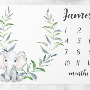 May include: A white baby milestone blanket with a watercolor illustration of a gray elephant with pink ears and a green leafy wreath. The blanket has the name "James" and numbers 1-12 for each month of the year.