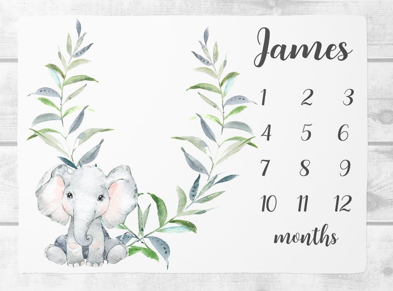 May include: A white baby milestone blanket with a watercolor illustration of a gray elephant with pink ears and a green leafy wreath. The blanket has the name "James" and numbers 1-12 for each month of the year.