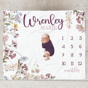 May include: A white baby blanket with a floral design and a baby girl sleeping on it. The blanket has a purple and pink floral border and the text "Wrenley Marie" and "months" printed on it. The numbers 1 through 12 are printed on the blanket.