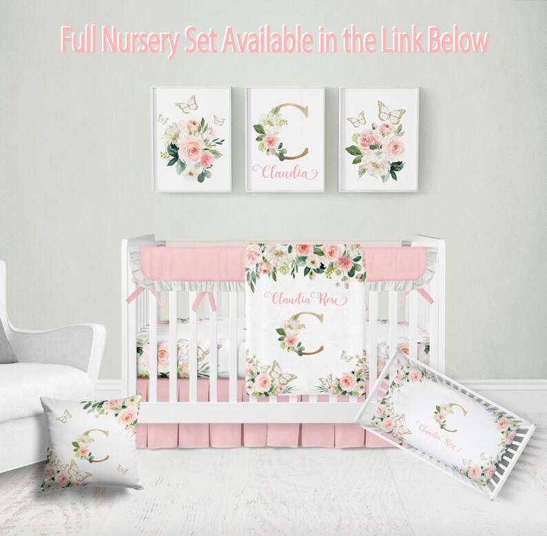 K&ouml;nnte beinhalten: Ein wei&szlig;es Babybett mit einem rosa-wei&szlig;en Blumenbettw&auml;sche-Set. Die Bettw&auml;sche umfasst einen Bettrock, eine Decke und zwei Kissen. Die Decke und die Kissen tragen den Namen "Claudia Rose" und einen goldenen Buchstaben C. Das Babybett ist mit drei gerahmten Drucken mit Schmetterlingen und Blumenmustern verziert.