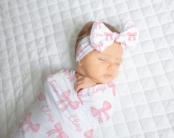 Könnte beinhalten: Ein Baby, eingewickelt in eine weiße Wickeldecke und mit einem passenden Stirnband, beide mit rosa Schleifen verziert. Der Name "Eloise" ist auf die Decke gedruckt. Das Baby liegt auf einer weißen, gesteppten Oberfläche.