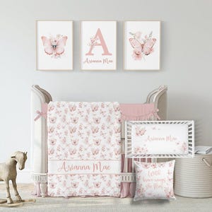 May include: A nursery set featuring a crib, bedding, and wall art. The set includes a crib, a blanket with a butterfly pattern, and three framed prints. The prints feature butterflies and the name "Arianna Mae". A decorative pillow with the text "With brave wings she flies" is also included.