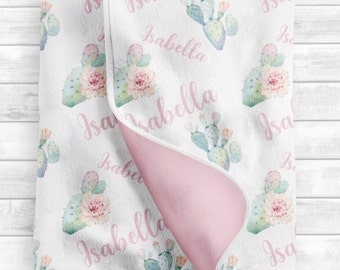 May include: A white baby blanket with a pink and green cactus print. The blanket is personalized with the name "Isabella" repeated throughout the design.