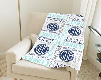May include: A white baby blanket with a repeating pattern of names and initials in navy blue, light blue, and gray. The blanket is draped over a beige armchair in a well-lit room.