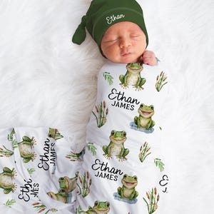 May include: A newborn baby wrapped in a white swaddle blanket and wearing a green hat. The blanket is decorated with cartoon frogs, cattails, and the name "Ethan James". The hat is green and has the name "Ethan" embroidered on it.