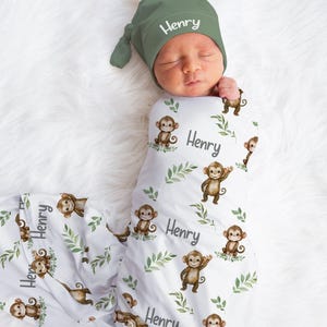 May include: A baby swaddled in a white blanket with a monkey and leaf pattern, and a matching green hat. The name "Henry" is printed on the hat and blanket. The blanket is wrapped around the baby.