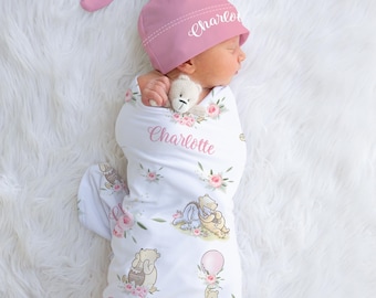 May include: A newborn baby wrapped in a white swaddle blanket and wearing a pink hat. The blanket features a floral and cartoon bear design with the name "Charlotte". The hat also has the name "Charlotte" embroidered on it. A small teddy bear is held by the baby.