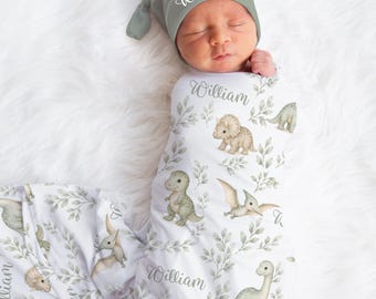 May include: A sage green knotted baby hat and a matching white swaddle blanket featuring a repeating pattern of green leaves and various dinosaurs. The name William is printed in a cursive script on both the hat and the swaddle fabric.