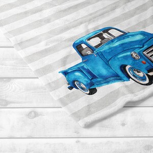 May include: A blue vintage truck illustration on a white and gray striped towel. The truck is a vibrant blue with white tires and chrome details. The towel is folded over a white wooden surface.