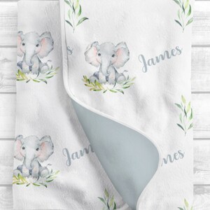 May include: A white baby blanket with a light blue backing. The blanket features a repeating pattern of watercolor illustrations of elephants with green leaves. The name "James" is written in cursive on the blanket.