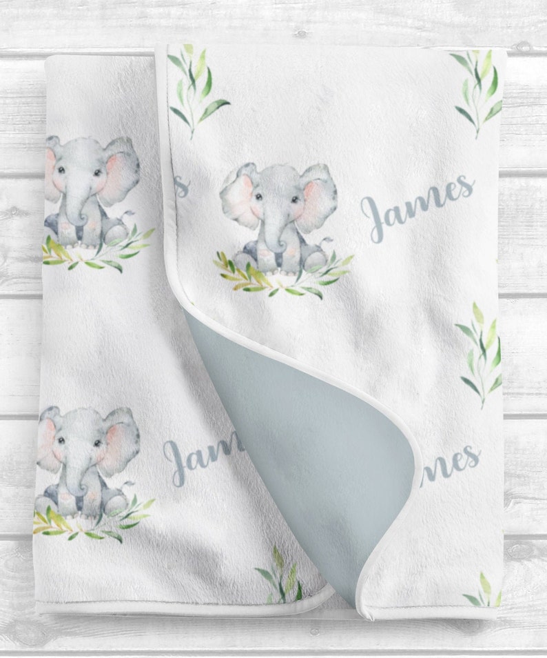 May include: A white baby blanket with a light blue backing. The blanket features a repeating pattern of watercolor illustrations of elephants with green leaves. The name "James" is written in cursive on the blanket.